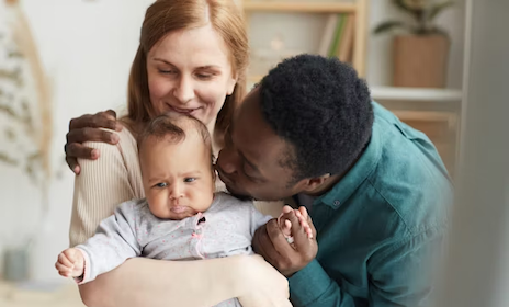 A happy interracial couple embraces their infant child in a warm, home setting, with the father gently kissing the baby while the mother looks on lovingly.